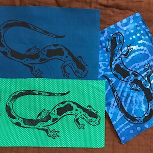 May include: Three fabric squares with a black and white salamander design printed on them. The squares are blue, green with white polka dots, and blue with a white swirl pattern.