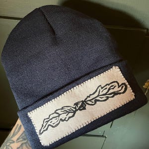 May include: A navy blue beanie with a white patch featuring a black line drawing of a leaf.