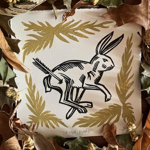 May include: A square white paper print featuring a black ink illustration of a running hare with an arrow through its body. The hare is framed by four gold-colored fern-like botanical prints. The text IRISH HARE is printed in black at the bottom.
