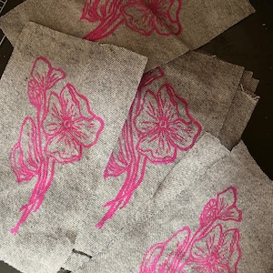 May include: Several gray fabric patches with a pink floral design. The flowers are stylized with detailed lines and are centered on the patches. The fabric has a textured appearance.