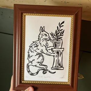 May include: Framed black and white print of a cat with a plant. The cat is sitting and holding the plant on a pedestal. The frame is brown with a gold inner border. The text "JUST DON'T" is at the bottom.
