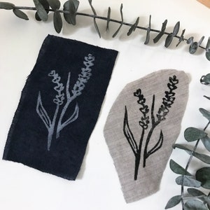 May include: Two fabric swatches with a black ink stamp of a simple lavender flower design. The fabric on the left is dark blue and the fabric on the right is a light beige.