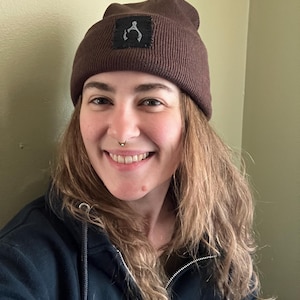 May include: A brown beanie with a square patch featuring a white graphic. The beanie is made of a ribbed knit material. The person in the image is wearing a dark blue zip-up hoodie.