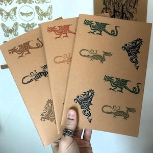 May include: Kraft paper notebooks with dragon illustrations in various colors, including red, green, and black. The notebooks feature different dragon designs. The image also shows butterfly and other illustrations.