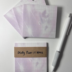 Puede incluir: Una pila de notas adhesivas de color lila pastel con un patrón de cuadrícula y un diseño de luna tenue. Las notas están etiquetadas como "Sticky Post-it Notes" en una etiqueta marrón.
