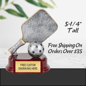 Puede incluir: Trofeo de pickleball con una pala y una pelota plateadas, sobre una base roja con una placa dorada para grabar. El trofeo mide 13,3 cm de alto. El texto "FREE CUSTOM ENGRAVING HERE" es visible.