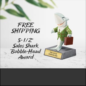 May include: A 5-1/2 inch tall sales shark bobblehead award. The shark is wearing a green plaid suit and holding a brown briefcase. The base of the award is gray with a gold plaque that says "Free Custom Engraving Here".