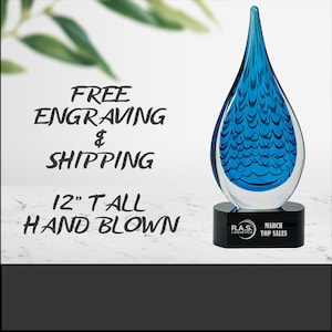 Op de afbeelding: Een handgeblazen glazen trofee, ongeveer 30 cm hoog, met een blauw druppelontwerp. De trofee staat op een zwarte voet met de tekst "March Top Sales". De afbeelding bevat ook de tekst "Free Engraving".