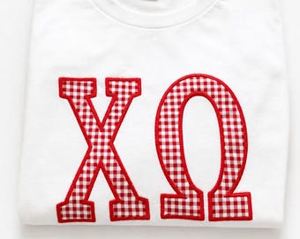 Puede incluir: Una camiseta blanca de manga corta con las letras griegas Chi y Omega aplicadas en la parte delantera. Las letras están rellenas con un patrón de cuadros vichy rojo y blanco y delineadas con un borde rojo grueso. Se ve una etiqueta negra.