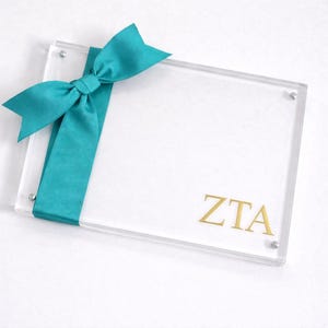 May include: Clear acrylic frame with a teal ribbon tied in a bow on the left side. The letters "ZTA" are printed in gold on the bottom right corner. The frame is held together with silver screws.