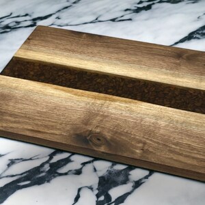 May include: A rectangular wooden chopping board with a brown wood grain. The board has a strip of clear resin embedded in the centre, filled with coffee beans.
