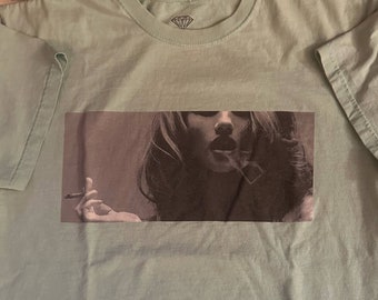 Puede incluir: Una camiseta verde claro con una imagen en blanco y negro de una mujer fumando un cigarrillo.