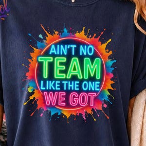 Może przedstawiać: Ciemnogranatowa koszulka z okrągłym wzorem przedstawiającym wielokolorowe plamy farby. Napis AIN'T NO TEAM LIKE THE ONE WE GOT wykonany jest jasnymi literami w stylu neonowym. Koszulka ma okrągły dekolt i krótkie rękawy.