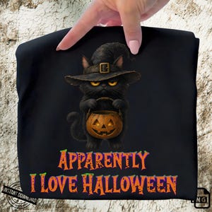 May include: A black t-shirt featuring a graphic of a black cat wearing a witch hat and holding a jack-o'-lantern pumpkin. Below the cat, the text reads "APPARENTLY I LOVE HALLOWEEN" in orange and purple letters with a distressed texture.