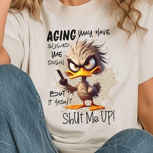 May include: White t-shirt with a cartoon duck and the text "Aging may have slowed me down, but it hasn't shut me up!"