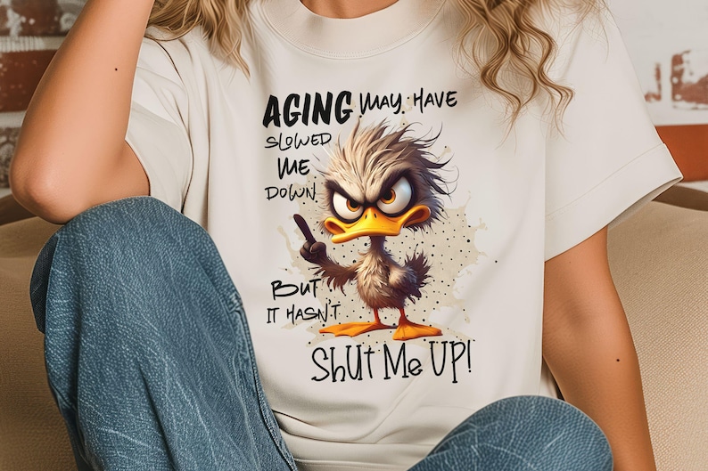 May include: White t-shirt with a cartoon duck and the text "Aging may have slowed me down, but it hasn't shut me up!"