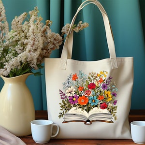 K&ouml;nnte beinhalten: Eine beige Tragetasche mit einem bunten Blumendesign, das auf der Vorderseite aufgedruckt ist. Die Blumen sind um ein offenes Buch mit sichtbaren Seiten angeordnet. Die Tasche hat zwei Henkel.