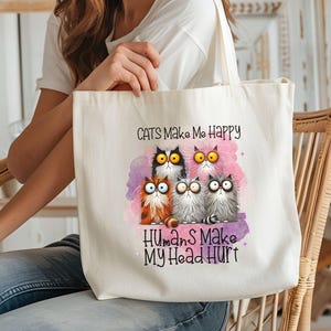 May include: A white canvas tote bag with a pink watercolor background and a group of cartoon cats. The text on the bag reads "Cats Make Me Happy Humans Make My Head Hurt."