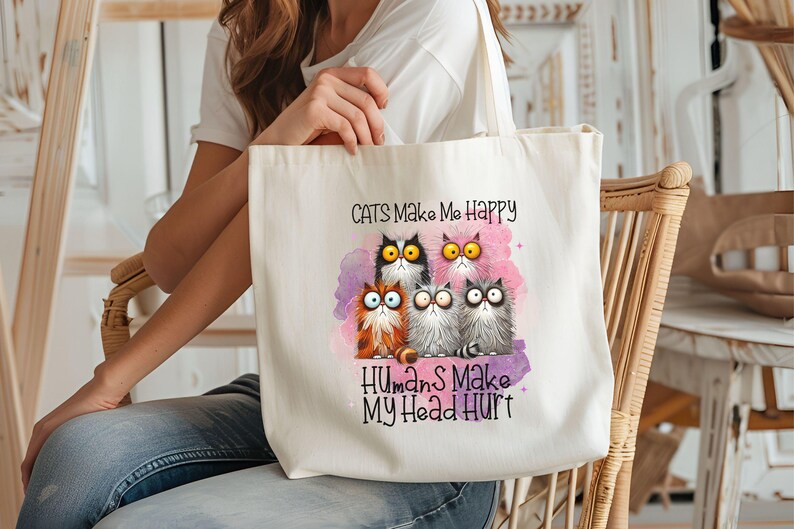 May include: A white canvas tote bag with a pink watercolor background and a group of cartoon cats. The text on the bag reads "Cats Make Me Happy Humans Make My Head Hurt."