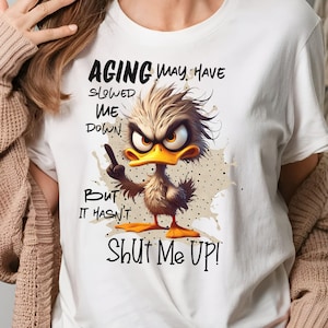 May include: White t-shirt with a cartoon duck and the text "Aging may have slowed me down, but it hasn't shut me up!"