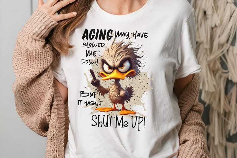 May include: White t-shirt with a cartoon duck and the text "Aging may have slowed me down, but it hasn't shut me up!"