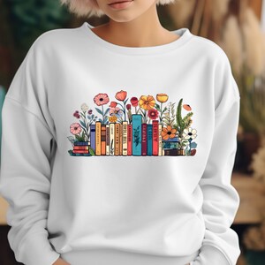 K&ouml;nnte beinhalten: Wei&szlig;er Pullover mit einem bunten Grafikdruck von einem Stapel B&uuml;cher mit Blumen, die darum herum wachsen.