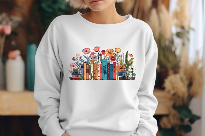 K&ouml;nnte beinhalten: Wei&szlig;er Pullover mit einem bunten Grafikdruck von einem Stapel B&uuml;cher mit Blumen, die darum herum wachsen.