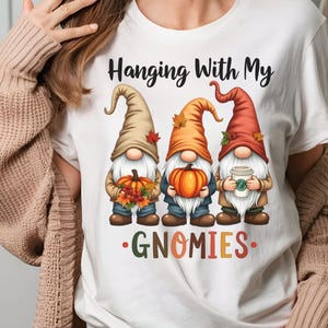 Könnte beinhalten: Weißes T-Shirt mit drei Zwergen, die Herbsthüte tragen und Kürbisse halten. Der Text "Hanging With My GNOMIES" ist oberhalb und unterhalb der Zwerge aufgedruckt. Ein Zwerg hält einen Kaffeebecher.