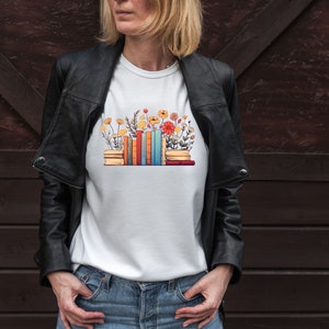 K&ouml;nnte beinhalten: Wei&szlig;es T-Shirt mit einem Aufdruck einer Stapel bunter B&uuml;cher, aus denen Blumen wachsen.
