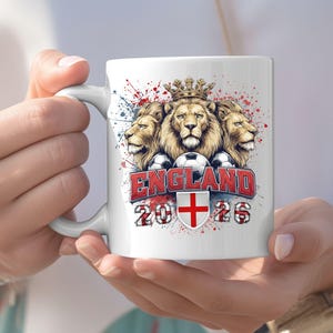 Puede incluir: Taza de cer&aacute;mica blanca con un dise&ntilde;o de tres leones coronados, balones de f&uacute;tbol y la palabra "ENGLAND" en rojo. La taza tambi&eacute;n muestra los a&ntilde;os "2026" y una cruz roja sobre un escudo blanco. El dise&ntilde;o tiene salpicaduras de pintura roja y azul.