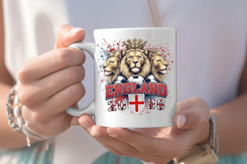 Puede incluir: Taza de cer&aacute;mica blanca con un dise&ntilde;o de tres leones coronados, balones de f&uacute;tbol y la palabra "ENGLAND" en rojo. La taza tambi&eacute;n muestra los a&ntilde;os "2026" y una cruz roja sobre un escudo blanco. El dise&ntilde;o tiene salpicaduras de pintura roja y azul.
