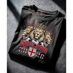 Puede incluir: Camiseta gris oscuro con un dise&ntilde;o de tres leones coronados, balones de f&uacute;tbol y el texto "ENGLAND 2026". El dise&ntilde;o incluye los colores de la bandera inglesa y un escudo.