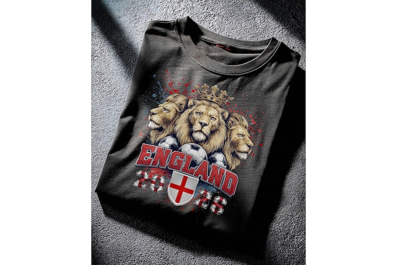 Puede incluir: Camiseta gris oscuro con un dise&ntilde;o de tres leones coronados, balones de f&uacute;tbol y el texto "ENGLAND 2026". El dise&ntilde;o incluye los colores de la bandera inglesa y un escudo.