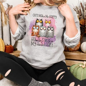 May include: A gray sweatshirt with a pink watercolor background and a group of cartoon cats. The text on the sweatshirt reads "Cats Make Me Happy Humans Make My Head Hurt".