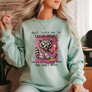 May include: A light blue sweatshirt with a graphic of a raccoon holding a beer and the text "don't invite me for 'casual drinks', there's nothing casual about the way I drink."