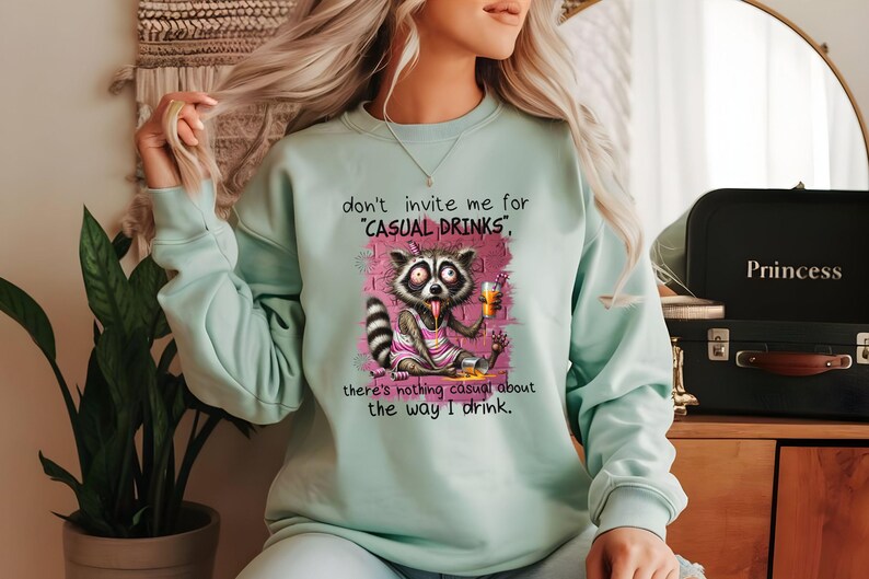May include: A light blue sweatshirt with a graphic of a raccoon holding a beer and the text "don't invite me for 'casual drinks', there's nothing casual about the way I drink."