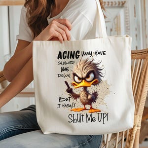 May include: White canvas tote bag with a cartoon duck illustration and the text "Aging may have slowed me down but it hasn't shut me up!"