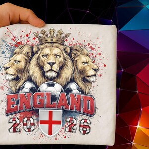 Puede incluir: Una camiseta color crema con un dise&ntilde;o que presenta tres leones coronados, balones de f&uacute;tbol y la palabra "ENGLAND" en rojo. El dise&ntilde;o incluye los a&ntilde;os "2026" y un escudo con una cruz roja.