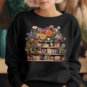 K&ouml;nnte beinhalten: Ein schwarzes Sweatshirt mit einem Aufdruck einer mit B&uuml;chern und Blumen gef&uuml;llten B&uuml;cherregals. Die Blumen sind in verschiedenen Farben, darunter Orange, Gelb, Rosa und Lila.