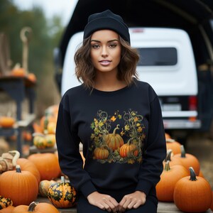 K&ouml;nnte beinhalten: Ein schwarzes Sweatshirt mit einem K&uuml;rbis-Design, das orange und gelbe Bl&auml;tter zeigt. Die Person tr&auml;gt eine schwarze M&uuml;tze. Der Hintergrund zeigt eine Ausstellung von K&uuml;rbissen, die ein herbstliches Erntethema hervorruft.