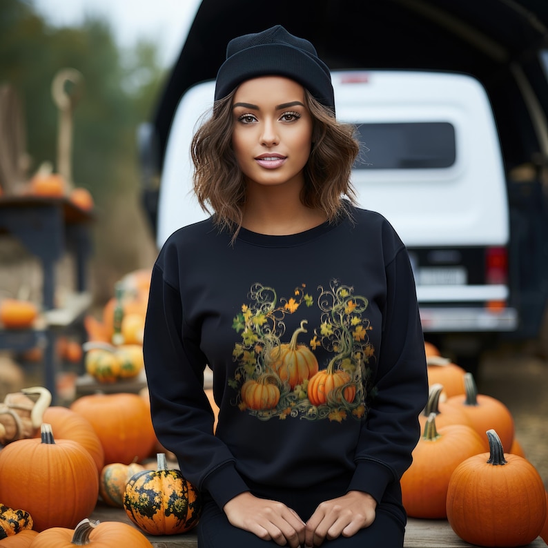 K&ouml;nnte beinhalten: Ein schwarzes Sweatshirt mit einem K&uuml;rbis-Design, das orange und gelbe Bl&auml;tter zeigt. Die Person tr&auml;gt eine schwarze M&uuml;tze. Der Hintergrund zeigt eine Ausstellung von K&uuml;rbissen, die ein herbstliches Erntethema hervorruft.