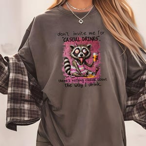 May include: A gray t-shirt with a raccoon cartoon wearing a pink dress and holding a glass of orange juice. The text on the shirt reads "don't invite me for 'CASUAL DRINKS', there's nothing casual about the way I drink."