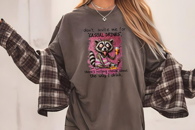 May include: A gray t-shirt with a raccoon cartoon wearing a pink dress and holding a glass of orange juice. The text on the shirt reads "don't invite me for 'CASUAL DRINKS', there's nothing casual about the way I drink."