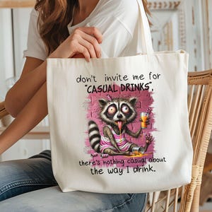 May include: White canvas tote bag with a cartoon raccoon drinking from a glass. The text on the bag reads "don't invite me for 'CASUAL DRINKS', there's nothing casual about the way I drink."