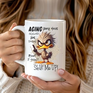 May include: White ceramic mug with a funny cartoon duck and the text "Aging may have slowed me down but it hasn't shut me up!"