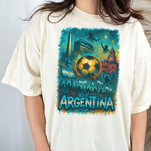 Puede incluir: Una camiseta blanca con un estampado gr&aacute;fico de un bal&oacute;n de f&uacute;tbol, banderas y la palabra "ARGENTINA" en un esquema de colores azul, verde y amarillo.
