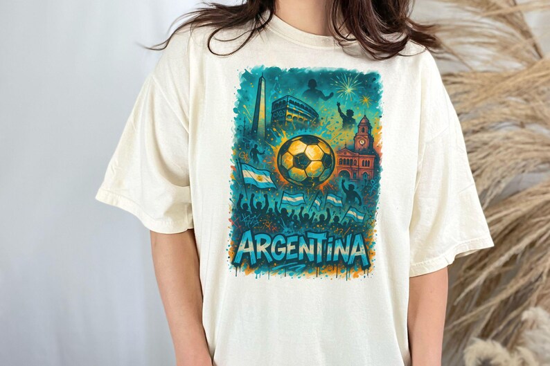Puede incluir: Una camiseta blanca con un estampado gr&aacute;fico de un bal&oacute;n de f&uacute;tbol, banderas y la palabra "ARGENTINA" en un esquema de colores azul, verde y amarillo.