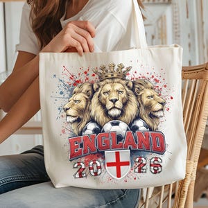 Puede incluir: Bolso tote color crema con un dise&ntilde;o de tres cabezas de le&oacute;n coronadas, balones de f&uacute;tbol y la palabra "ENGLAND" en rojo. La bolsa tambi&eacute;n muestra el a&ntilde;o "2026" y la bandera inglesa. El dise&ntilde;o tiene salpicaduras de pintura rojas y azules.