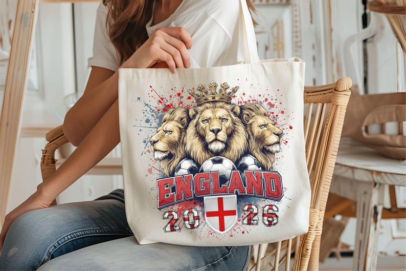 Puede incluir: Bolso tote color crema con un dise&ntilde;o de tres cabezas de le&oacute;n coronadas, balones de f&uacute;tbol y la palabra "ENGLAND" en rojo. La bolsa tambi&eacute;n muestra el a&ntilde;o "2026" y la bandera inglesa. El dise&ntilde;o tiene salpicaduras de pintura rojas y azules.