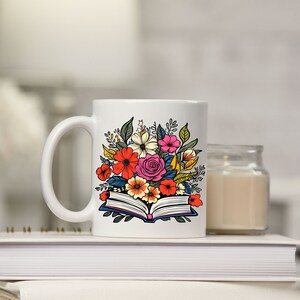 K&ouml;nnte beinhalten: Wei&szlig;e Keramiktasse mit einem bunten Blumenmuster auf einem offenen Buch. Die Tasse steht auf einem Stapel B&uuml;cher.